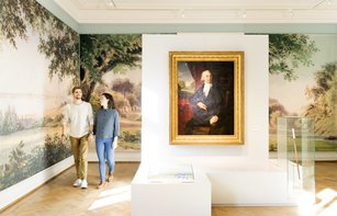 Besuchende sind im Ausstellungsraum zu sehen. Die Wand ist mit einer Landschaftstapete versehen