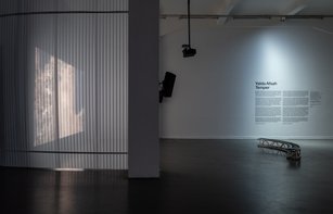 Ein großer abgedunkelter Raum, in dem links durch eine halbtransparente Wand eine Filmprojektion zu sehen ist. Rechts ist ein Text an der Wand geplättet und eine gebogene Metallbank zu sehen.