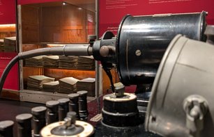 Technisches Gerät mit Trafo (Pantostat) im Vordergrund, im Hintergrund eine rote Ausstellungswand mit Vitrine, darin historische Krankenakten.