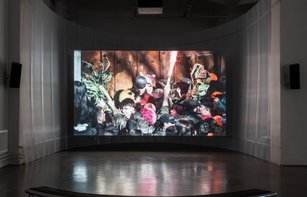 In einem großen, abgedunkelten Raum hängt eine große Leinwand, auf der ein Film gezeigt wird. Dieser zeigt eine gedrängte Menschenmenge. Eine Person hält einen funkensprühenden Feuerwerkskörper nach oben.
