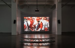 In einem großen, abgedunkelten Raum hängt zwischen zwei Säulen eine große Leinwand, auf der ein Film gezeigt wird. Dieser zeigt eine Menschenmenge. Alle Personen sind ähnlich gekleidet. Die Person im Mittelpunkt trägt Handschuhe und hält Feuerwerkskörper in der Hand. 