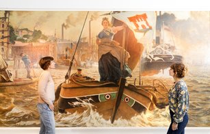 Zwei Menschen schauen sich das große Gemälde der Altonia an. Sie ist eine Frau, die auf einem Segelschiff steht. Im Hintergrund sind Dampfschiffe und der Hafen zu sehen.