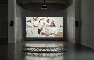 In einem großen, abgedunkelten Raum hängt eine große Leinwand, auf der ein Film gezeigt wird. Dieser zeigt in weiß gekleidete Menschen, die sich an den Händen halten und einen Tanz aufführen. 